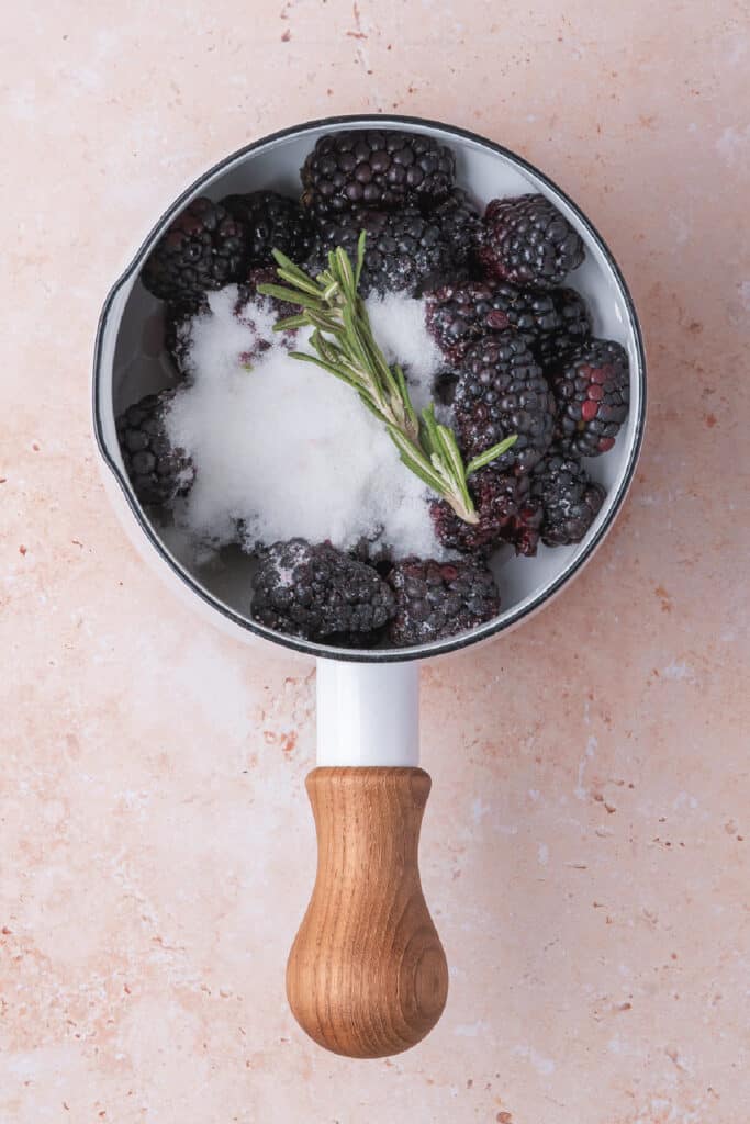 Fresh blackberries, rosemary, and sugar in a white saucepan before cooking the jam
