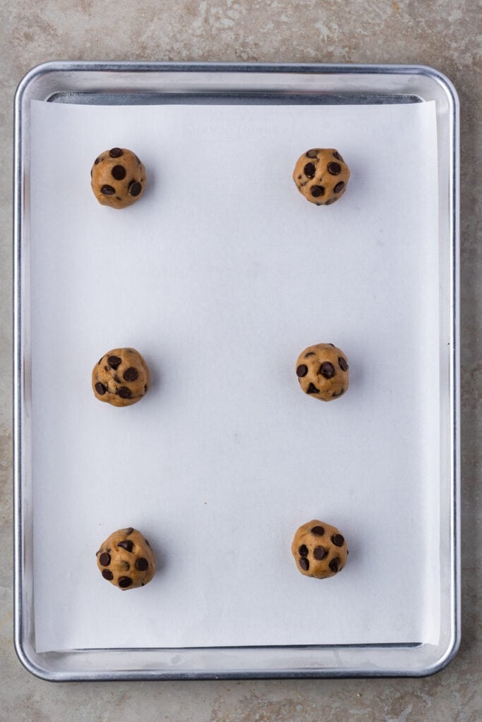 Six scoops of chocolate chip cookie dough evenly spaced on a parchment-lined baking sheet.