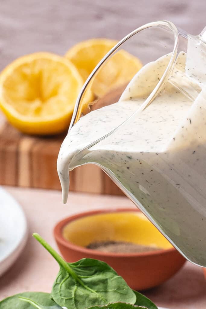 Pouring creamy lemon dill dressing from a glass pitcher over fresh spinach.