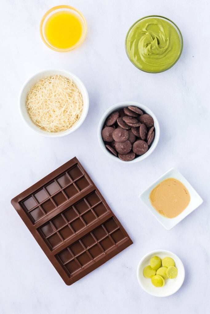 Flat lay of ingredients for Dubai Chocolate Bar including kataifi pastry, pistachio butter, tahini, melting chocolate, melted butter, lime green candy melts, and a silicone chocolate bar mold.