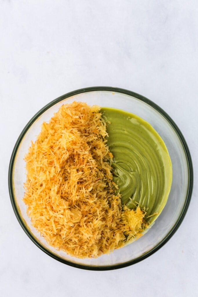 Toasted kataifi pastry being folded into pistachio-tahini mixture in a glass mixing bowl.