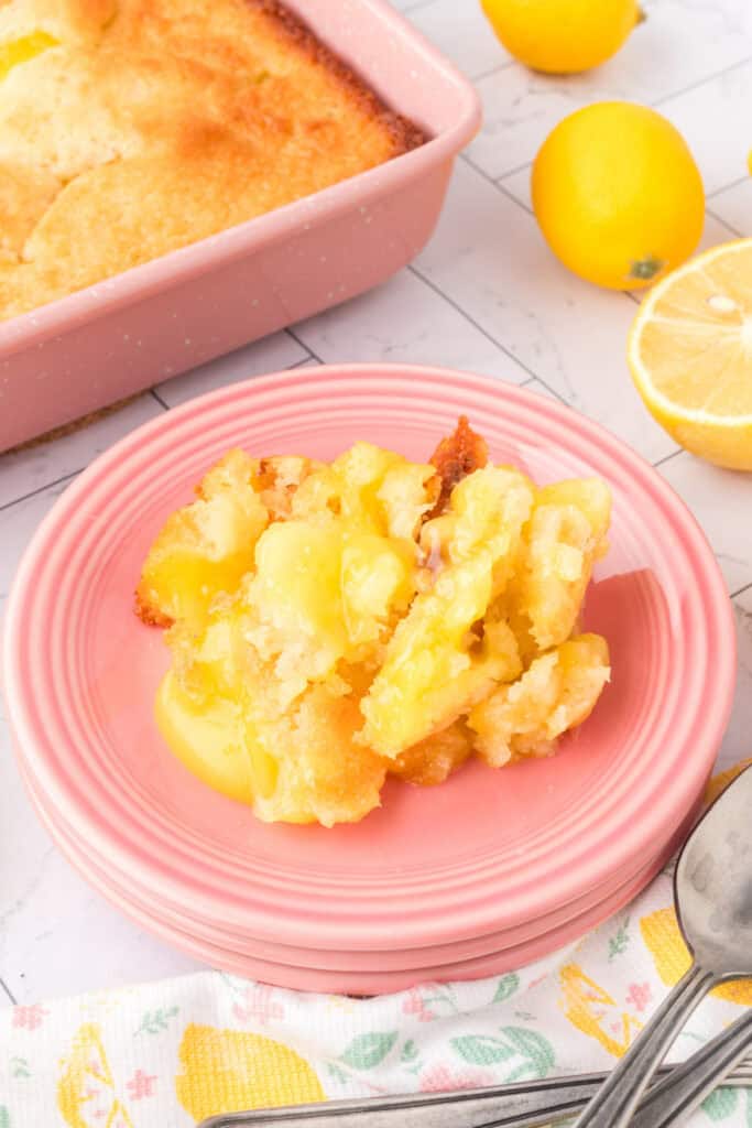 Plate of Easy Lemon Cobbler in front of the baking dish, surrounded by lemons and a lemon-printed towel