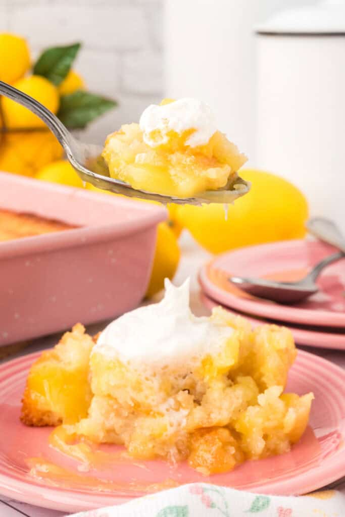 Spoonful of Easy Lemon Cobbler with whipped topping held above a plated serving, with lemons in the background