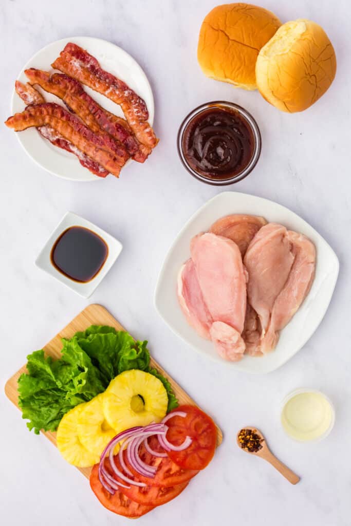 Ingredients for grilled Hawaiian chicken sandwich including bacon, buns, BBQ sauce, chicken breasts, soy sauce, lettuce, pineapple, tomato, and red onion.