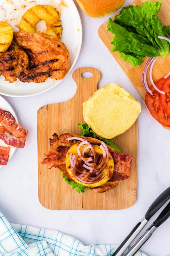 Grilled Hawaiian chicken sandwich layered with bacon, pineapple, red onion, and BBQ sauce on a cutting board.