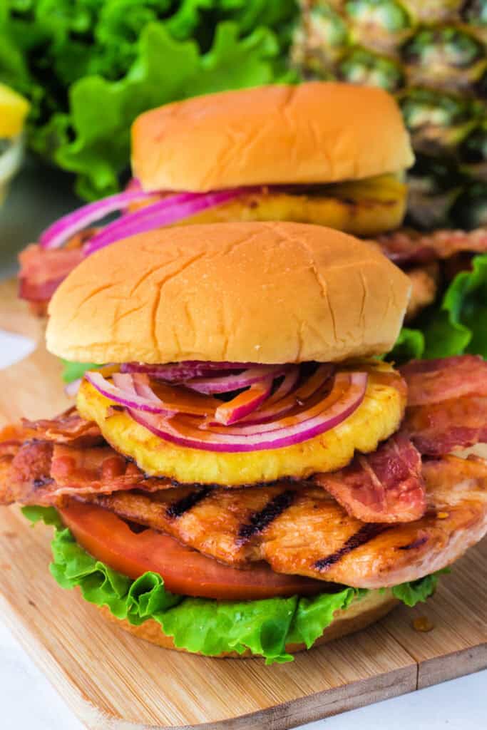 Close-up of a stacked grilled Hawaiian chicken sandwich showing layers of chicken, bacon, pineapple, and red onion.