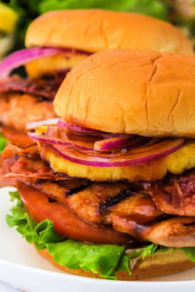 Side view of two grilled Hawaiian chicken sandwiches stacked with bacon, pineapple, lettuce, and tomato.