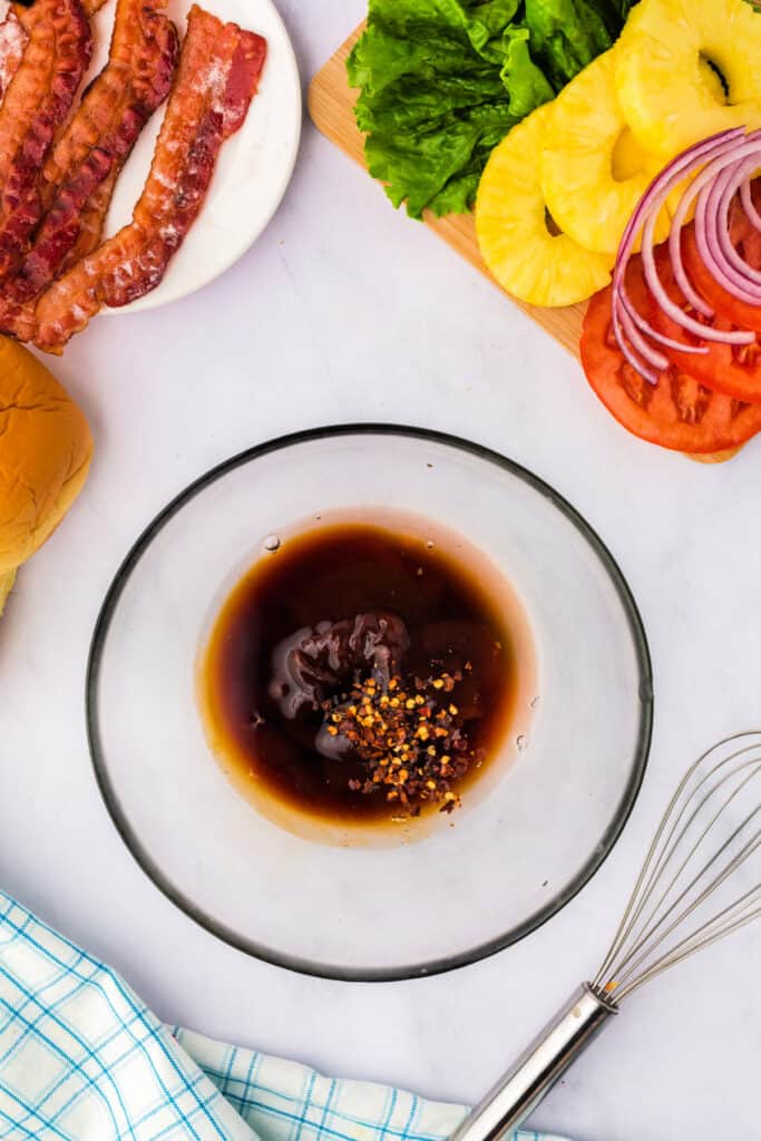 Overhead view of marinade ingredients in a bowl surrounded by bacon, lettuce, pineapple, tomato, and sandwich buns.