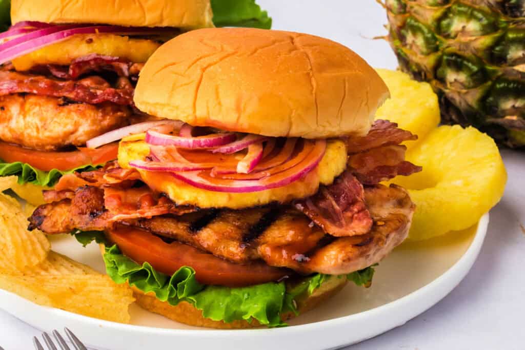 Close-up of grilled Hawaiian chicken sandwiches served on a plate with fresh pineapple slices and potato chips.