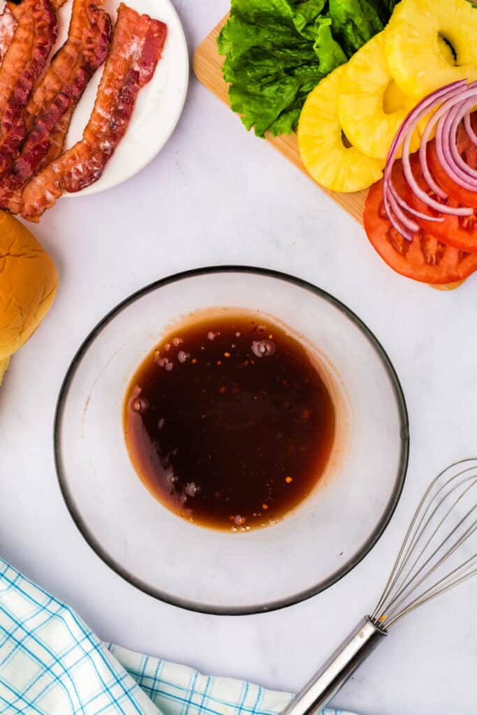 Marinade mixed together in a glass bowl with bacon, lettuce, pineapple, tomato, and buns nearby.
