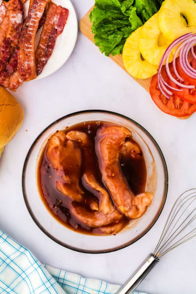 Chicken breasts marinating in the pineapple BBQ sauce mixture with surrounding sandwich ingredients.
