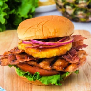 Square format image of a grilled Hawaiian chicken sandwich piled high with lettuce, tomato, bacon, pineapple, and red onion on a soft bun.