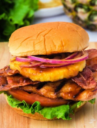Square format image of a grilled Hawaiian chicken sandwich piled high with lettuce, tomato, bacon, pineapple, and red onion on a soft bun.