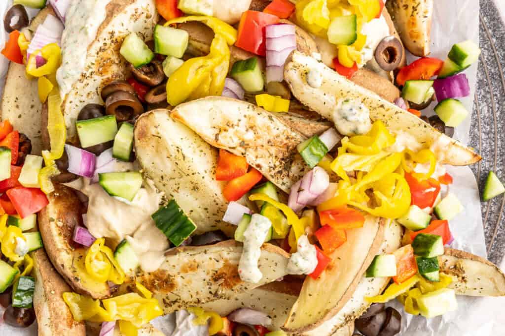 Horizontal close-up of loaded potato wedges with black olives, banana peppers, cucumbers, and diced peppers.