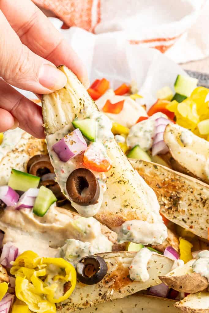 Close-up of a hand holding a seasoned potato wedge topped with yogurt sauce, olives, and diced veggies.