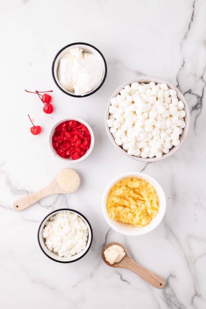 Overhead view of individual ingredients in small bowls, including mini marshmallows, cottage cheese, whipped topping, crushed pineapple, chopped maraschino cherries, sugar, and mayonnaise.