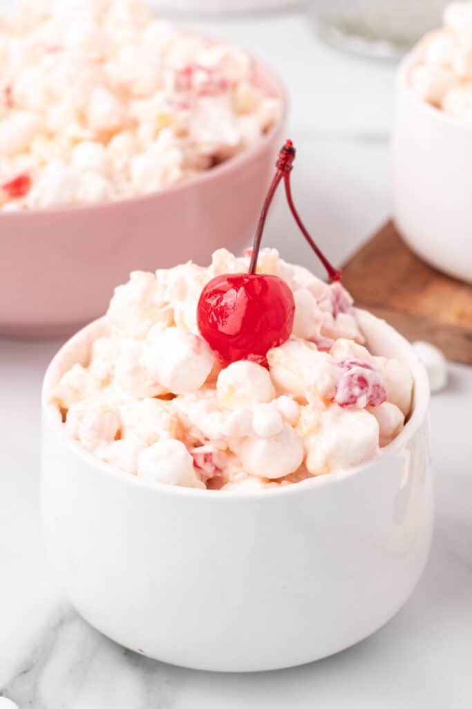 Close-up of fruit salad served in a white bowl, topped with a maraschino cherry.