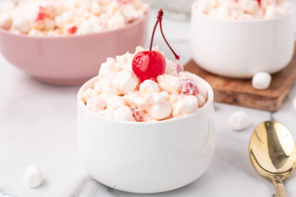 White bowl of million dollar fruit salad in focus, garnished with a cherry, with additional bowls in the background.