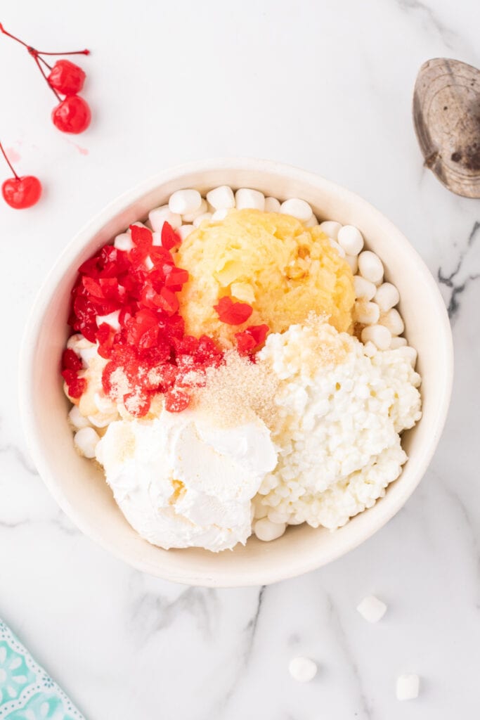 Ingredients for fruit salad layered in a large mixing bowl, ready to be combined - includes marshmallows, pineapple, cherries, whipped topping, sugar, cottage cheese, and mayonnaise.