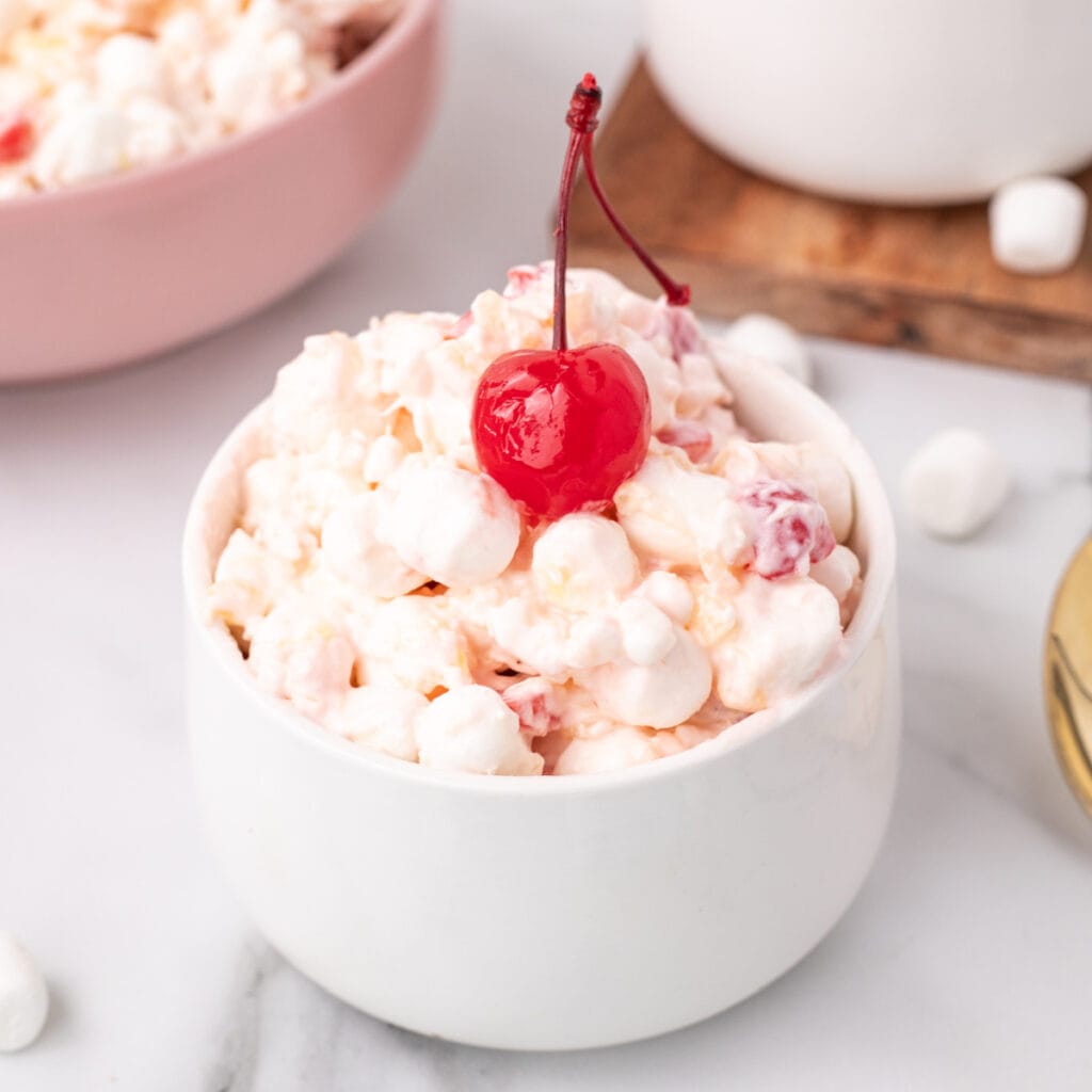 Square-format image of a white bowl filled with fluffy fruit salad, garnished with a maraschino cherry, perfect for social media sharing.