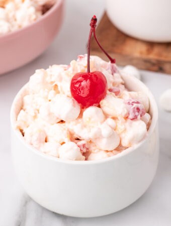 Square-format image of a white bowl filled with fluffy fruit salad, garnished with a maraschino cherry, perfect for social media sharing.
