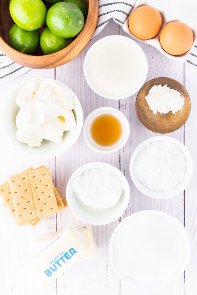Overhead view of mini key lime cheesecake ingredients including limes, graham crackers, butter, eggs, and cream cheese
