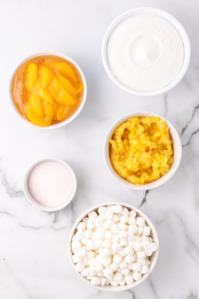 Overhead view of ingredients for peach fluff salad in bowls on a marble surface.