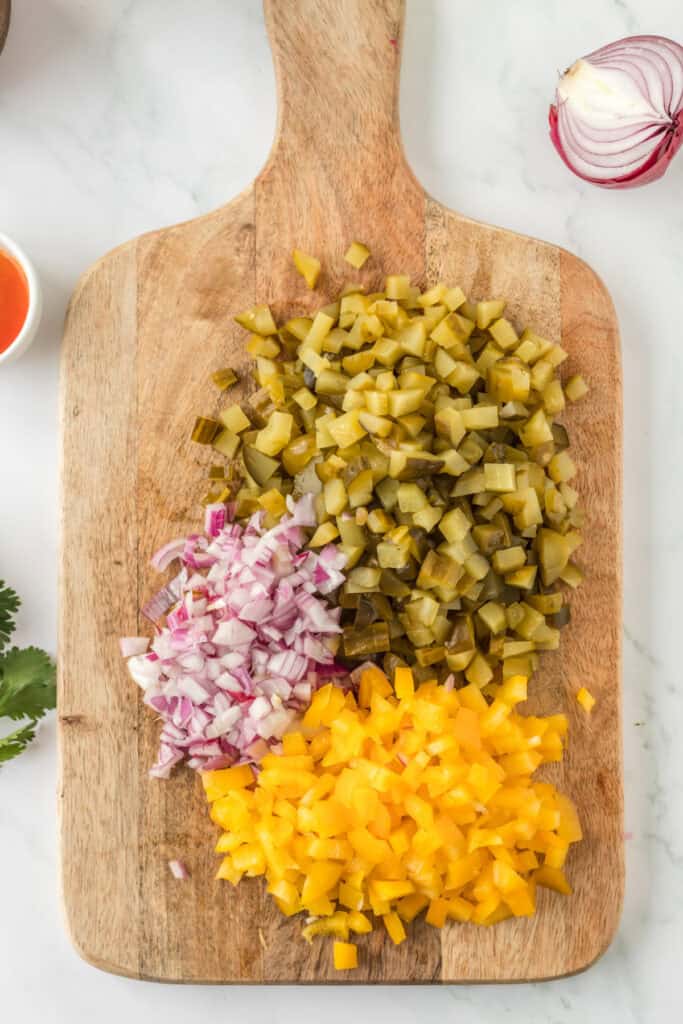 Chopped dill pickles, red onion, and yellow bell pepper on a wooden cutting board, ready for mixing.