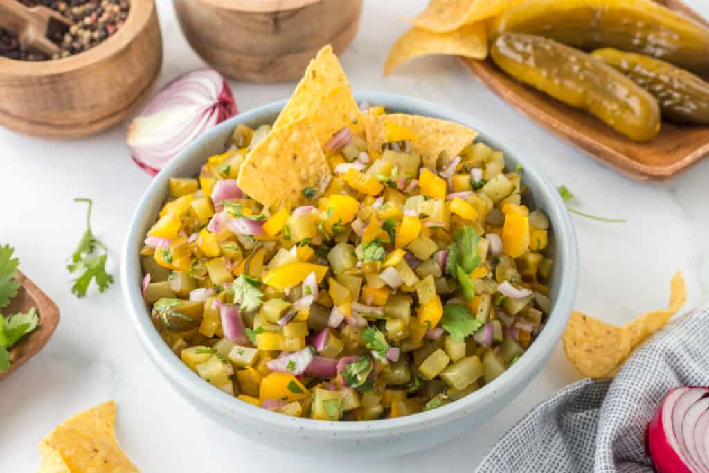A bowl of Pickle de Gallo with tortilla chips, surrounded by ingredients like red onion, pickles, and spices.