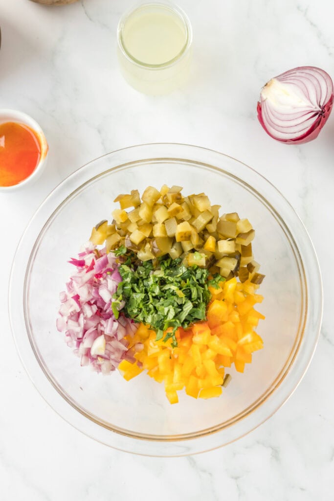 Ingredients for Pickle de Gallo in a glass mixing bowl, including chopped pickles, red onion, yellow bell pepper, and fresh cilantro.