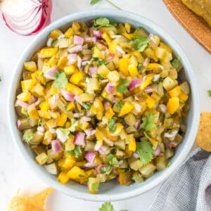 Square-cropped overhead shot of Pickle de Gallo in a bowl, surrounded by tortilla chips and fresh herbs.
