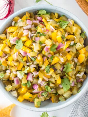 Square-cropped overhead shot of Pickle de Gallo in a bowl, surrounded by tortilla chips and fresh herbs.