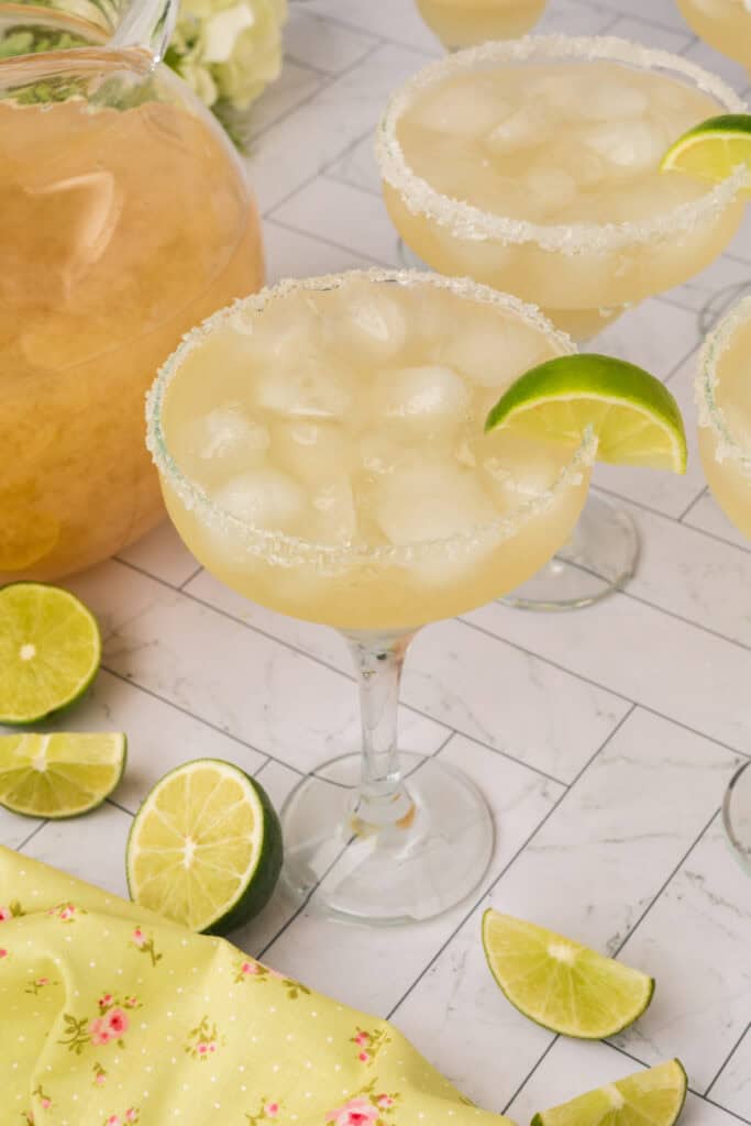Salt-rimmed margarita glass garnished with a lime wedge and filled with ice next to a pitcher