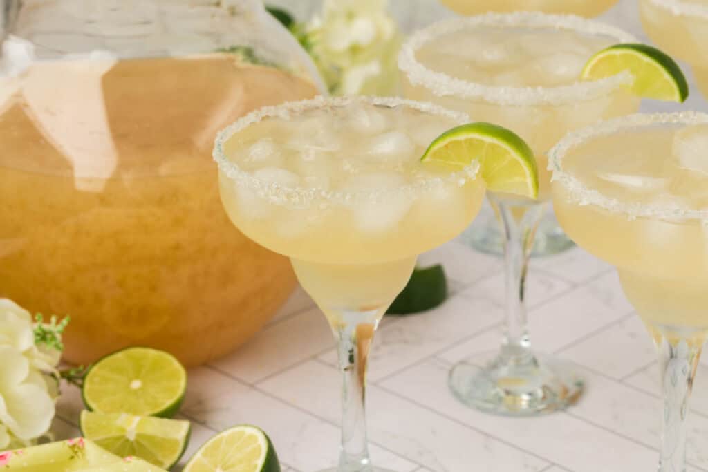Several margarita glasses filled with ice and garnished with lime, with a pitcher in the background