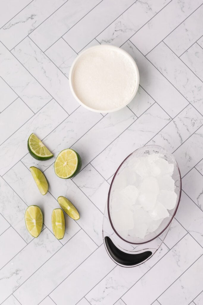 Ice, lime wedges, and a bowl of salt arranged on a tiled surface for preparing margaritas