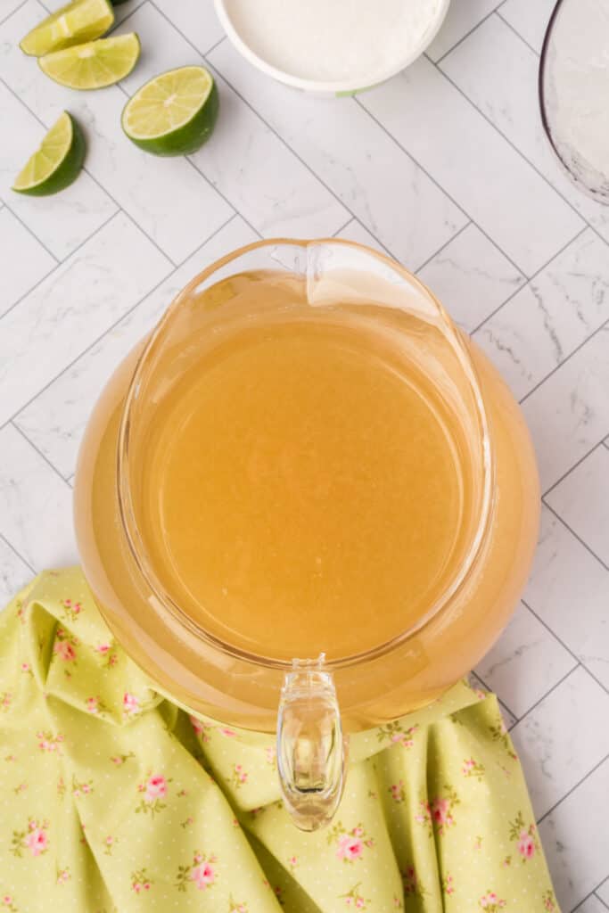 Large glass pitcher filled with margarita mix surrounded by lime wedges and salt