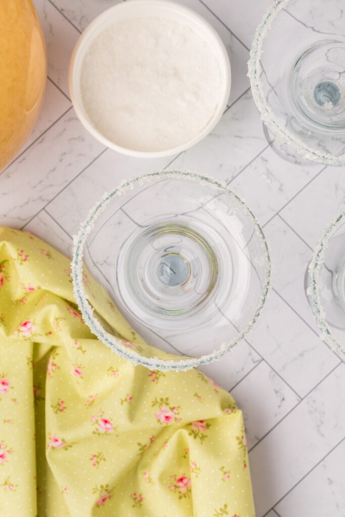 Close-up of margarita glasses rimmed with salt next to a bowl of salt and floral fabric