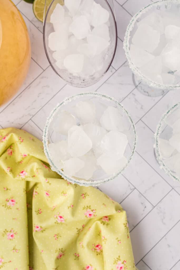 Salt-rimmed margarita glasses filled with ice next to a pitcher of mix and lime wedges