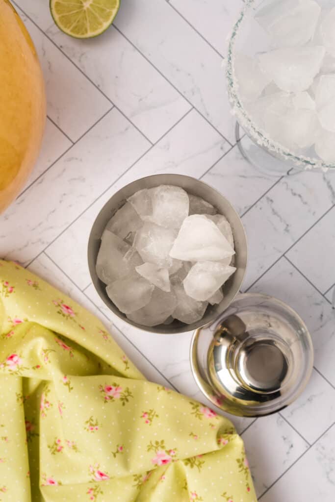 Cocktail shaker filled with ice next to a margarita glass and a pitcher of mix