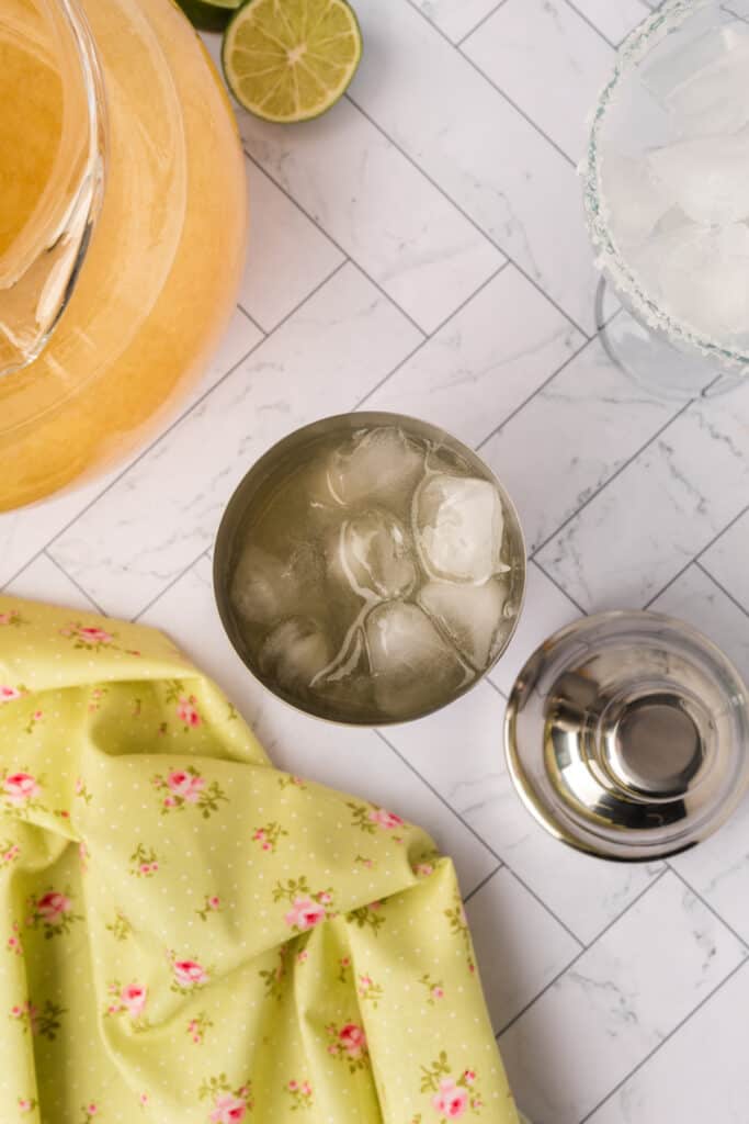 Margarita mix and ice in a shaker ready to be poured, with limes and pitcher nearby