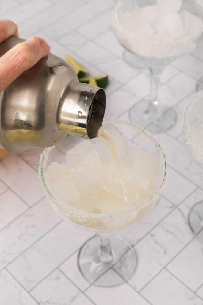 Margarita being poured from a cocktail shaker into a salt-rimmed glass with ice