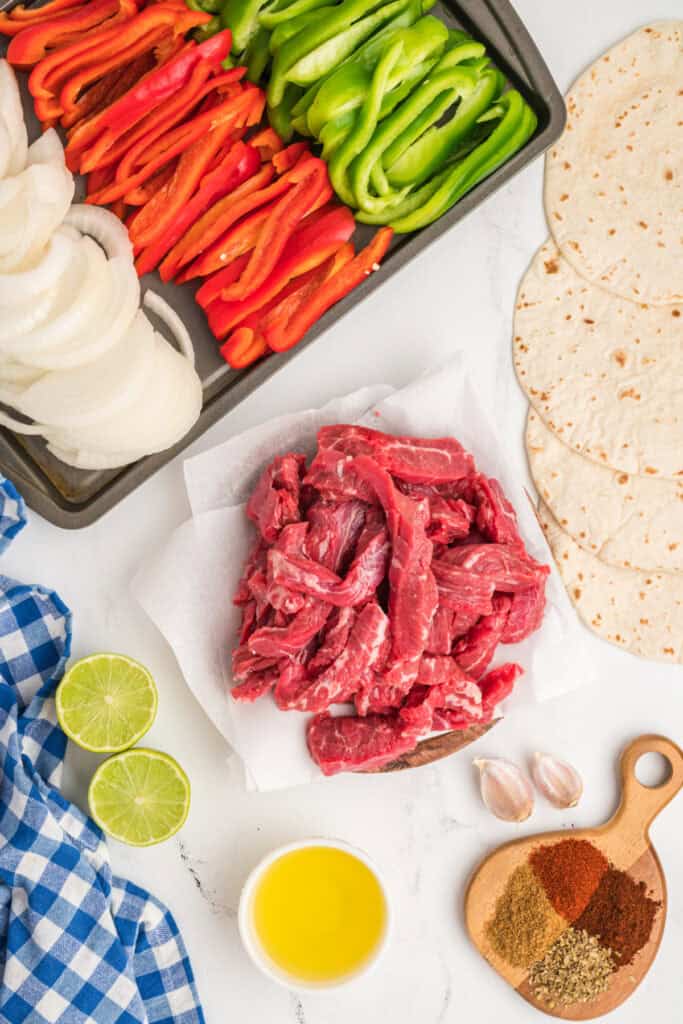 Overhead view of raw fajita ingredients including sliced peppers, onions, flank steak, lime, garlic, olive oil, and spices.