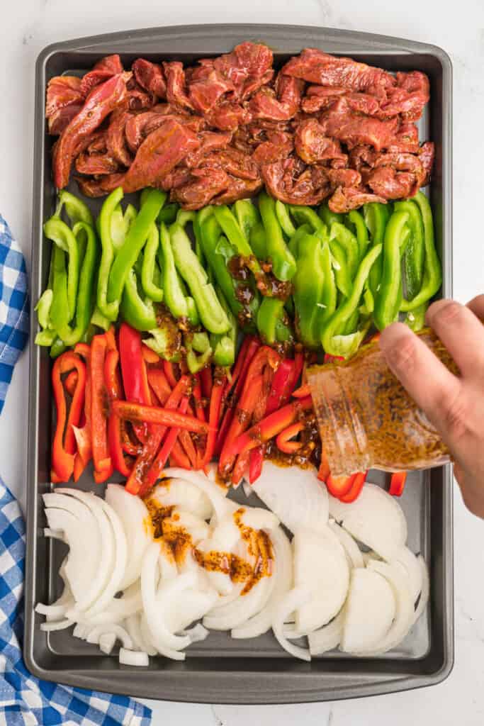 Raw steak and sliced peppers and onions arranged on a sheet pan with marinade being poured over.