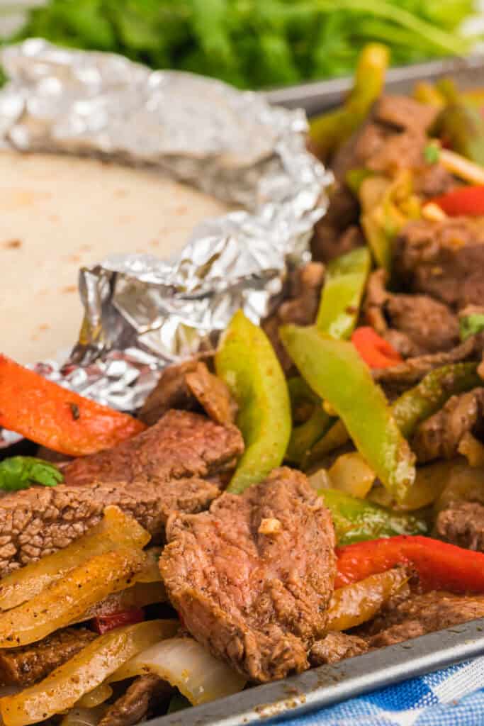 Sheet pan of cooked steak fajitas with foil-wrapped tortillas in the corner and fresh herbs in the background.
