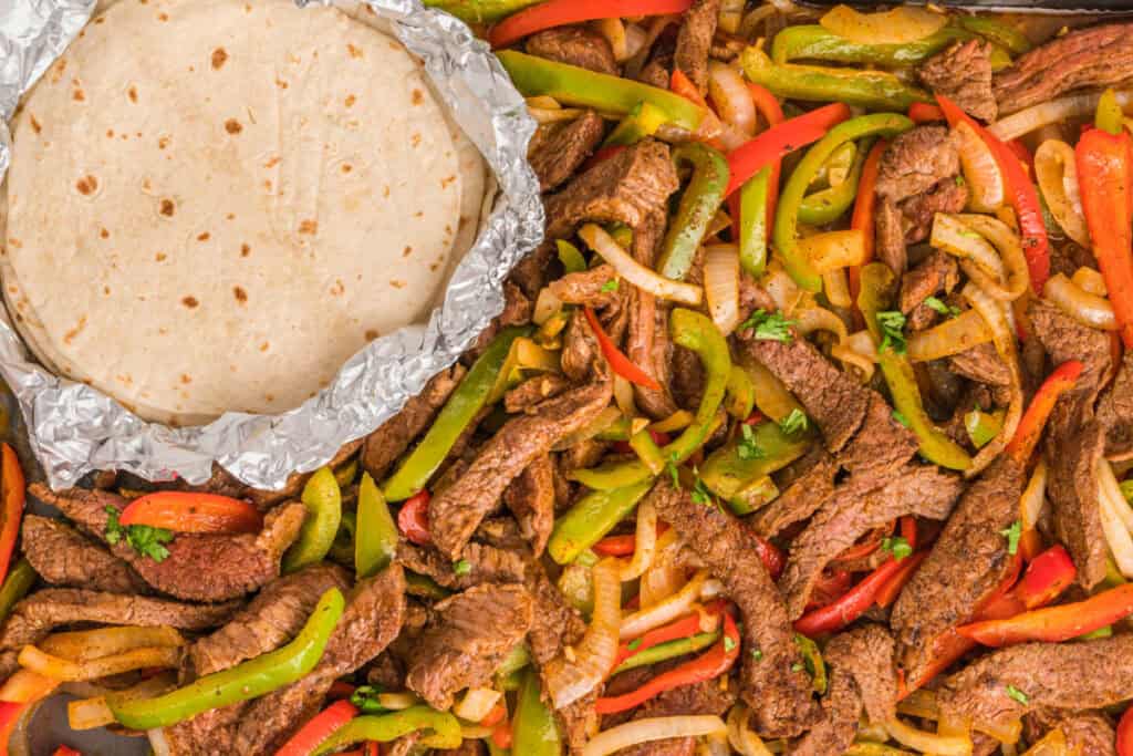 Overhead shot of cooked steak, onions, and bell peppers next to a stack of soft tortillas wrapped in foil.