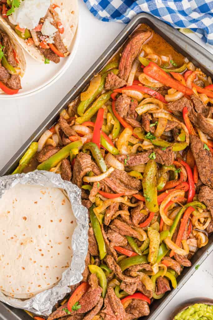 Full sheet pan of cooked steak fajitas with tortillas in foil and a partial plate of tacos in the corner.