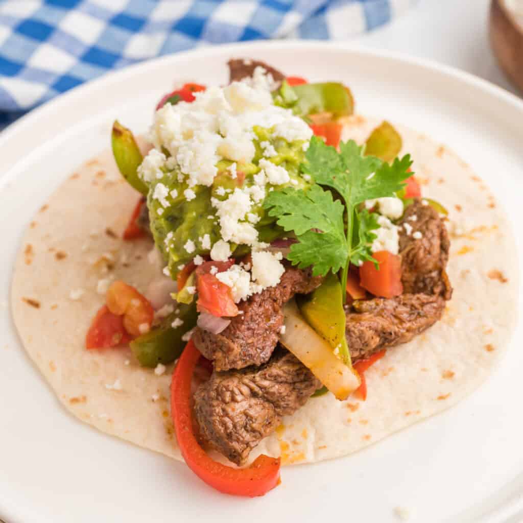 Close-up square image of a steak fajita taco with guacamole, cheese, and cilantro on a white plate.