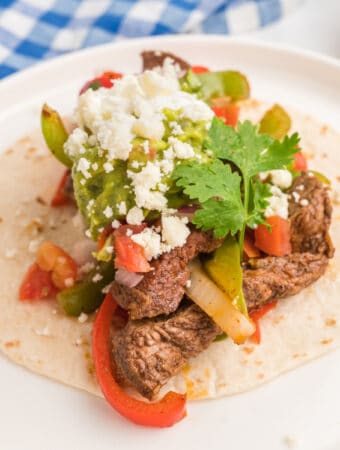 Close-up square image of a steak fajita taco with guacamole, cheese, and cilantro on a white plate.