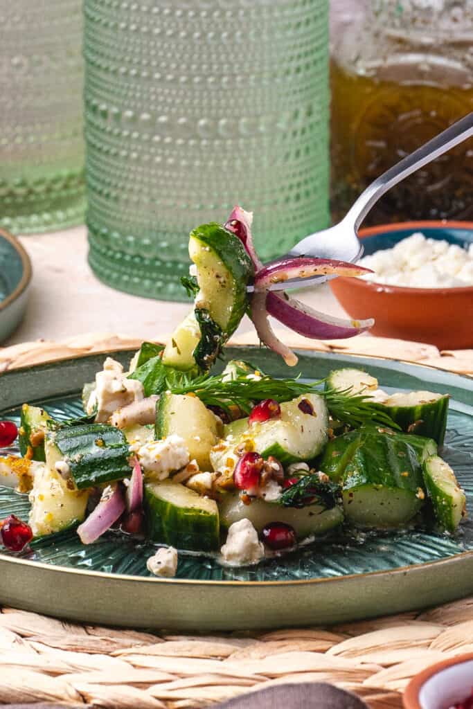 Close-up of fork lifting cucumber salad with red onion, feta, and herbs from a green plate