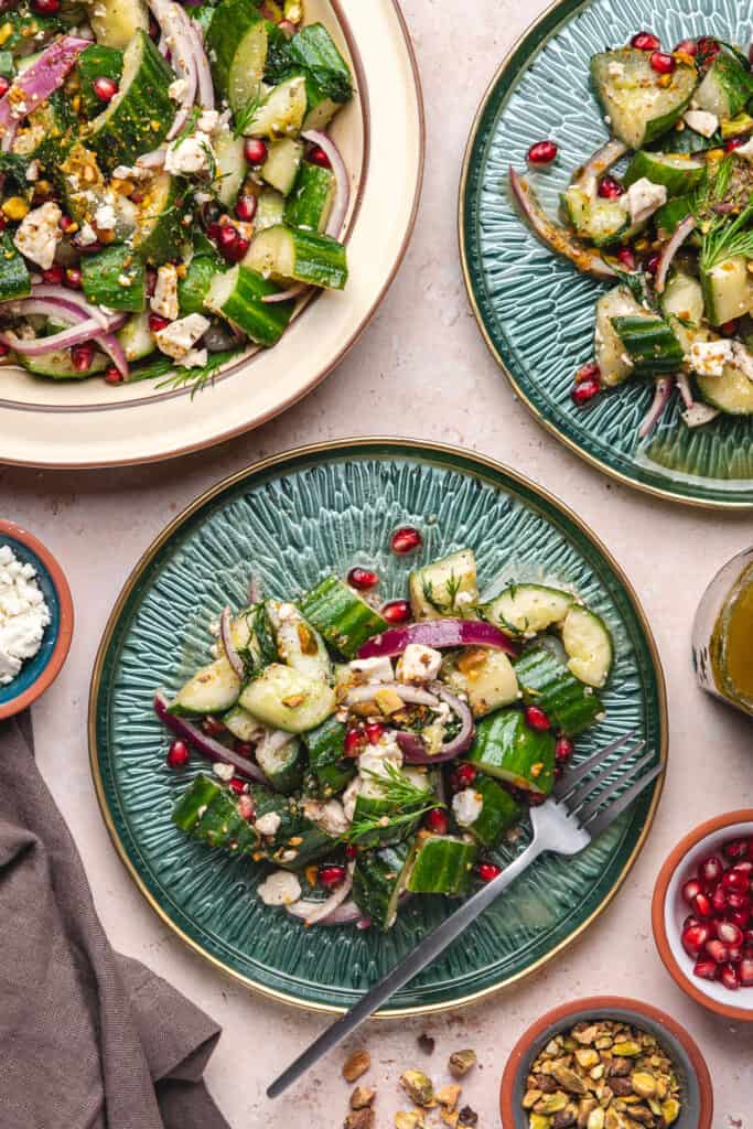 Overhead view of three green plates and serving platter with portions of cucumber salad, surrounded by toppings